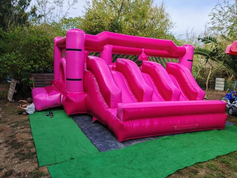 Bright pink inflatable bounce house with multiple slide lanes on green mat in outdoor garden setting