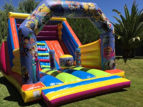 Colorful inflatable bounce house with slide and landing pad on grass, featuring bright primary colors and cartoon character designs